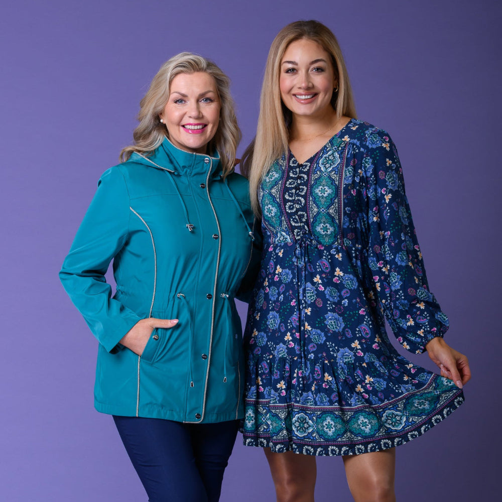 Two women modeling apparel against a solid purple studio background. The woman on the left wears a teal hooded jacket, navy cropped pants, and white sneakers.